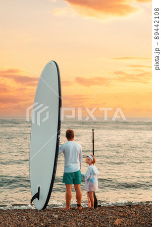 Summer surfing. Father and daughter standing at the beach with a sup board. Ocean and the sunset in the background. Back view. Vertical Summer surfing. Father and daughter standing at the beach with a sup board. Ocean and the sunset in the background. Back view. Vertical 89442108