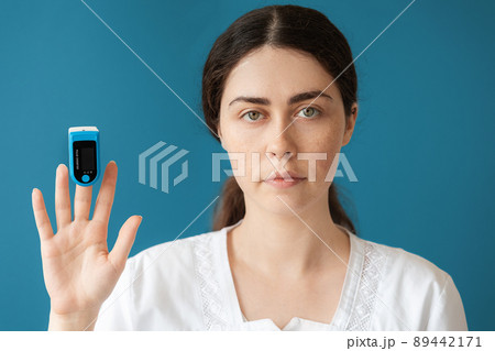 Portrait of woman doctor shows a pulse oximeter on her index finger. Blue background. The concept of check health and medical insurance Portrait of woman doctor shows a pulse oximeter on her index finger. Blue background. The concept of check health and medical insurance 89442171