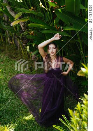 A girl in an evening long dress stands by a banana thicket posing for a photographer 89452836