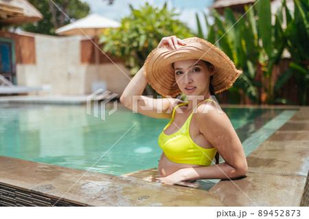 A girl in a sun hat and swimsuit in the water pool leaned her elbow on the edge of the parapet and poses for a photo against a backdrop of tropical plants 89452873
