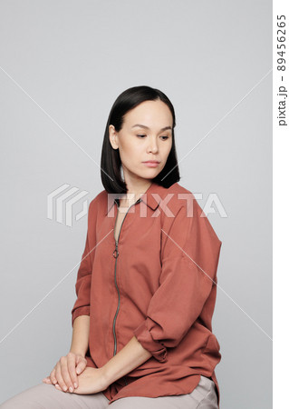 Sad young Asian woman in dark beige shirt sitting on stool and looking down Sad young Asian woman in dark beige shirt sitting on stool and looking down 89456265
