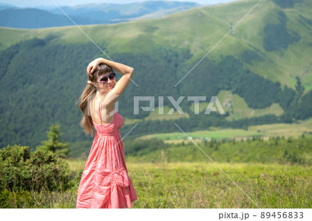 Young woman in red dress standing on grassy field on a windy day in summer mountains enjoying view of nature. 89456833
