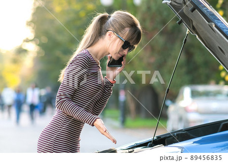 Young upset woman driver talking on mobile phone near a broken car with open hood waiting for help having trouble with her vehicle on a city street. 89456835