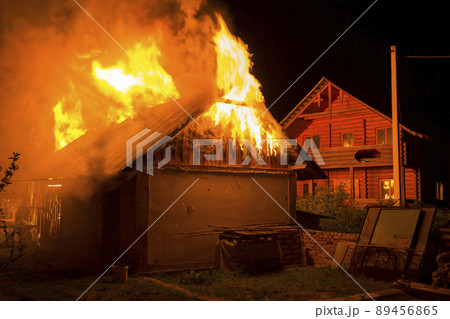 Wooden barn burning at night. High orange fire flames, dense smoke from under tiled roof on dark sky, trees silhouettes and residential wooden neighbor cottage background. Disaster and danger concept. 89456865