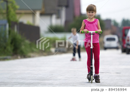 Pretty young long-haired blond child girl in pink clothing with scooter on sunny suburb street and boy on skateboard blurred summer bright background. Children activities, games and fun concept. Pretty young long-haired blond child girl in pink clothing with scooter on sunny suburb street and boy on skateboard blurred summer bright background. Children activities, games and fun concept. 89456994