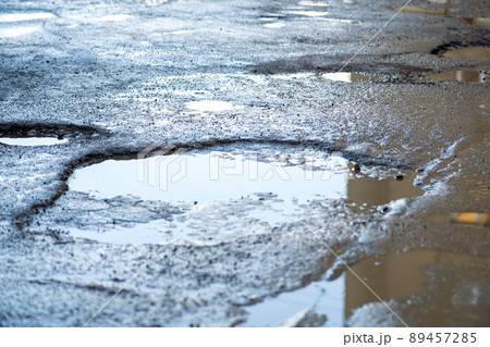 Close up of a road in very bad condition with big potholes full of dirty rain water pools. 89457285