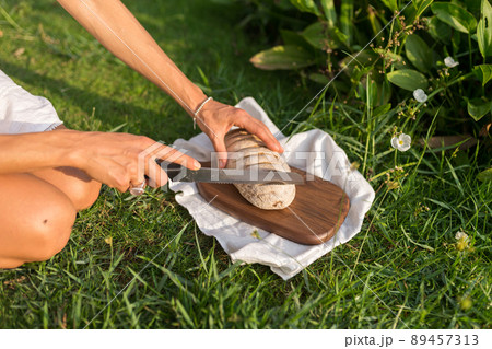 Composition of homemade white flour bread on wood board with white cloth napkin on the green grass hand cutting bread with a knife outdoors.  89457313