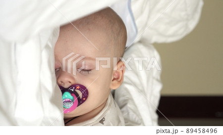 Vertical close-up face newborn chubby sleeping baby lying on stomach open eyes and smile covered with blanket on bed on white background. infancy, childhood, development, medicine and health concept Vertical close-up face newborn chubby sleeping baby lying on stomach open eyes and smile covered with blanket on bed on white background. infancy, childhood, development, medicine and health concept 89458496