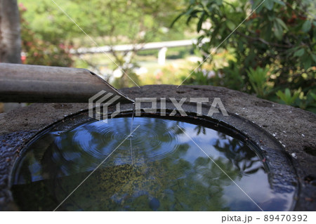 蹲、つくばいのアップ、竹筒から滴り落ちる水が手水鉢の表面に輪紋を作り、水鏡に空や樹々の葉が写る、横 89470392