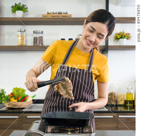 Young housewife using BBQ Clamp Shovel Roasting the steak. Morning atmosphere in a modern kitchen. Young housewife using BBQ Clamp Shovel Roasting the steak. Morning atmosphere in a modern kitchen. 89476850