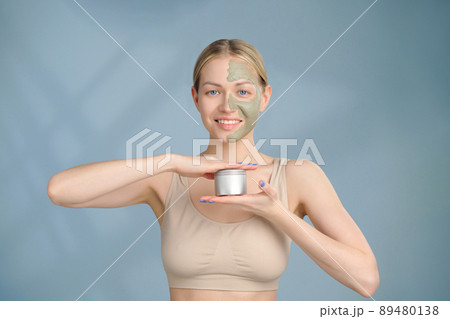 A close-up face portrait of a young blonde woman. Green beauty mask on half of the face. A woman holds a jar of moisturizer in her hands. Blue background. 89480138