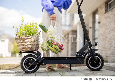 Woman drives on electrical scooter with mesh bag full of vegetables, close-up Woman drives on electrical scooter with mesh bag full of vegetables, close-up 89485315