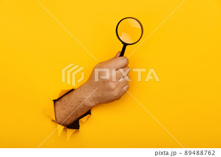 Hand holds black magnifying glass in torn hole of yellow background. Searching information concept Hand holds black magnifying glass in torn hole of yellow background. Searching information concept 89488762