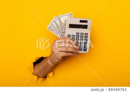 Male hand with calculator and dollar banknotes through a paper hole in yellow background Male hand with calculator and dollar banknotes through a paper hole in yellow background 89488763