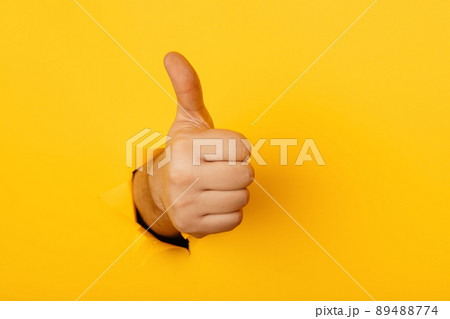 Hand shows a thumb up through a hole of yellow paper wall 89488774
