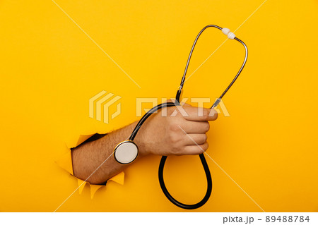Hand holds a stethoscope out of a hole torn in yellow paper wall with copy space Hand holds a stethoscope out of a hole torn in yellow paper wall with copy space 89488784