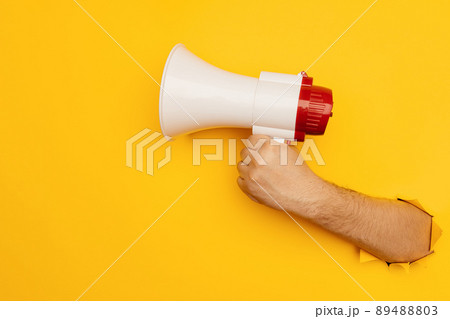 Hand holds a megaphone from a hole in the wall on a yellow background with copy space Hand holds a megaphone from a hole in the wall on a yellow background with copy space 89488803