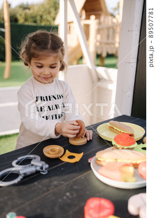 Happy little girl playing on toy kitchen on wheels. Cute girl make a burger on toy kitchen outdoors. Adorable kid play on fresh air 89507781