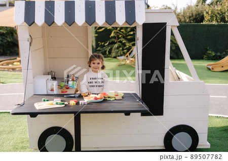 Happy little girl playing on toy kitchen on wheels. Cute girl make a burger on toy kitchen outdoors. Adorable kid play on fresh air Happy little girl playing on toy kitchen on wheels. Cute girl make a burger on toy kitchen outdoors. Adorable kid play on fresh air 89507782