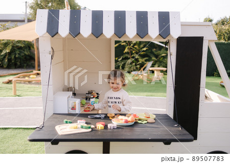 Happy little girl playing on toy kitchen on wheels. Cute girl make a burger on toy kitchen outdoors. Adorable kid play on fresh air Happy little girl playing on toy kitchen on wheels. Cute girl make a burger on toy kitchen outdoors. Adorable kid play on fresh air 89507783