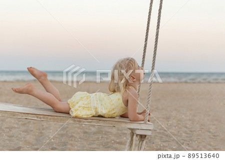 Little caucasian girl rides rope swing on sea background. Cute blonde toddler on the beach at sunset. Little caucasian girl rides rope swing on sea background. Cute blonde toddler on the beach at sunset. 89513640