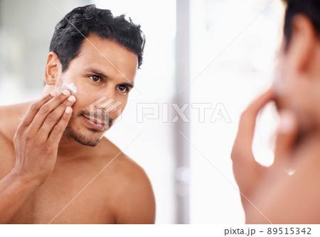He takes great care of his skin. A young man applying cream to his face while looking in the mirror. 89515342
