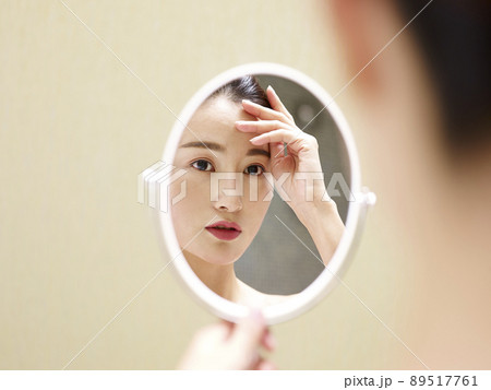 beautiful young asian woman looking at self in mirror, hand on forehead 89517761