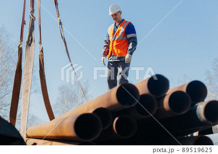 Slinger in helmet unloads metal pipes on construction site on sunny summer day. Real scene. Workflow Slinger in helmet unloads metal pipes on construction site on sunny summer day. Real scene. Workflow 89519625