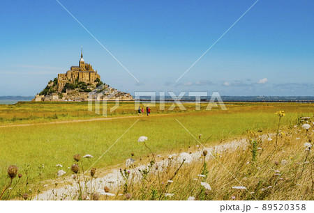 Tourists crossing empty green fields to visit the famous Le Mont Saint Michel tidal island on a bright sunny day, Normandy, Northern France Tourists crossing empty green fields to visit the famous Le Mont Saint Michel tidal island on a bright sunny day, Normandy, Northern France 89520358