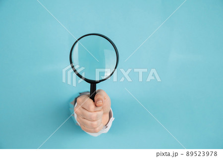 A woman's hand sticking out of a hole in a paper blue background holds a magnifying loupe. Copy space.  89523978