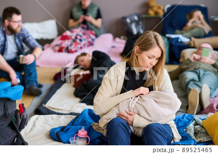 Young blond female refugee lulling baby wrapped into warm plaid on bed 89525465