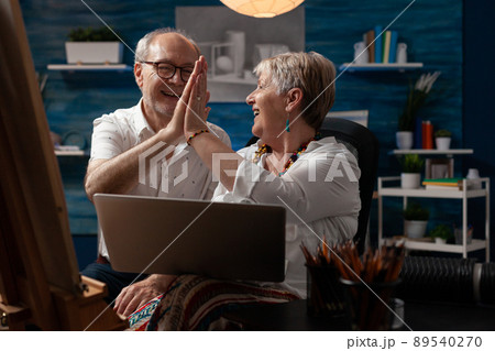 Elderly artist couple holding laptop doing high five hand gesture celebrating selling artwork in online auction sitting in home studio. Senior art creators enjoying success on social media. 89540270