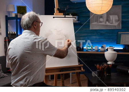 Senior man feeling inspired looking at ceramic vase doing creative pencil drawing on easel sitting down in professional art studio. Elderly artist in the evening shading black and white sketch. Senior man feeling inspired looking at ceramic vase doing creative pencil drawing on easel sitting down in professional art studio. Elderly artist in the evening shading black and white sketch. 89540302