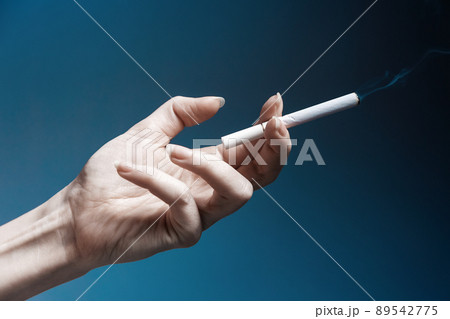 World No Tabacco day. A pale, crooked female hand, close-up, holding a smoking cigarette. Dark blue background. The concept of nicotine addiction World No Tabacco day. A pale, crooked female hand, close-up, holding a smoking cigarette. Dark blue background. The concept of nicotine addiction 89542775