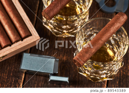 View of a wooden box with Cuban cigars, a lighter and a cutter. Still life with two glasses of whiskey. View of a wooden box with Cuban cigars, a lighter and a cutter. Still life with two glasses of whiskey. 89543389
