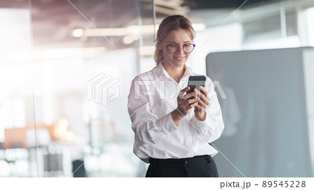Businesswoman wearing glasses with mobile phone in hand standing in modern office 89545228