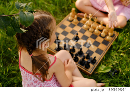 Two little girls are playing chess on a wooden chessboard outdoors in nature. One of the girls teaches the game and the rules of another child. Close-up. Rear view Two little girls are playing chess on a wooden chessboard outdoors in nature. One of the girls teaches the game and the rules of another child. Close-up. Rear view 89549344