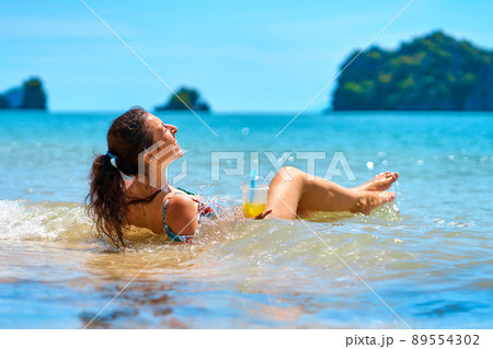 A beautiful slender girl in a swimsuit enjoys a tan while lying on a tropical beach, holding a glass of pineapple juice in her hand. A beautiful slender girl in a swimsuit enjoys a tan while lying on a tropical beach, holding a glass of pineapple juice in her hand. 89554302
