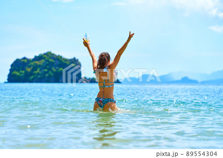 A beautiful slender girl in a swimsuit enjoys dancing in the ocean, holding a glass of pineapple fresh in her hand. A beautiful slender girl in a swimsuit enjoys dancing in the ocean, holding a glass of pineapple fresh in her hand. 89554304