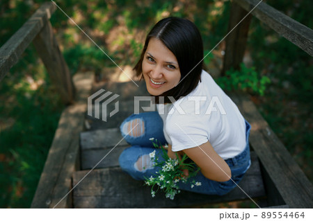A laughing woman is sitting in the park on the old wooden stairs with a bouquet of white wild flowers. 89554464