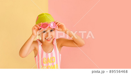 Funny joy girl in striped swimsuit and a pool cap, puts on swimming glasses, isolated on an pink and yellow studio background. Learning to swim in the pool from an early age. Happy baby Funny joy girl in striped swimsuit and a pool cap, puts on swimming glasses, isolated on an pink and yellow studio background. Learning to swim in the pool from an early age. Happy baby 89556468