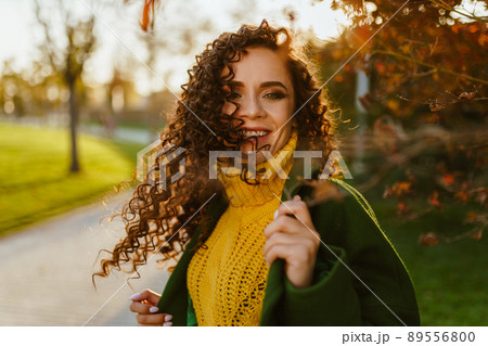 a girl with curly brown hair and a charming smile circling her head so that her hair fluttered in the wind and smiles holding on to the lapel of her coat in the park in the autumn 89556800