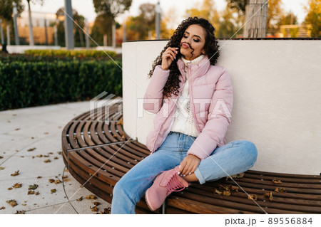 On a wooden bench sits tucking under one leg a beautiful girl and plays the curl of her long curly hair applying it over the upper lip 89556884