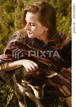 On the steppe grass sits a girl dressed in the style of hippies or boh and strokes the dog 89556887