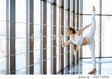 Attractive girl in white sportswear stretches her legs near the panoramic window Attractive girl in white sportswear stretches her legs near the panoramic window 89574099