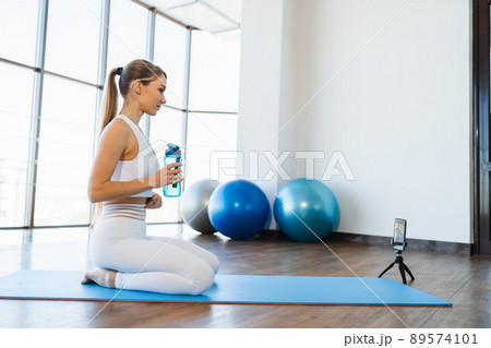Young fitness woman sitting on yoga mat with bottle of water and live streaming workout 89574101