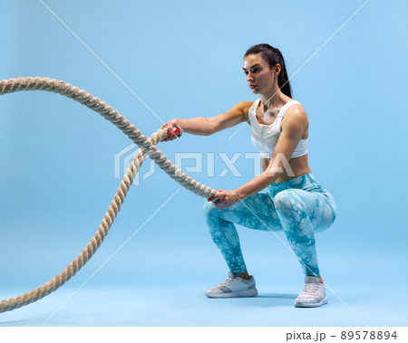 Dynamic portrait of young sportive woman training, doing exercises with rope isolated over blue studio background Dynamic portrait of young sportive woman training, doing exercises with rope isolated over blue studio background 89578894