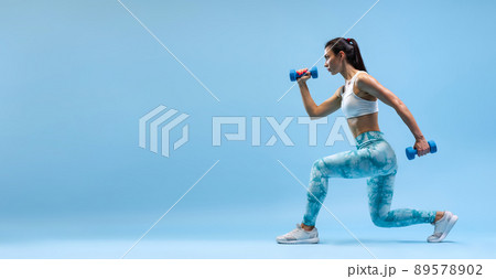 Side view portrait of young sportive girl training, doing leg exercises with dumbbell isolated over blue studio background. Flyer 89578902