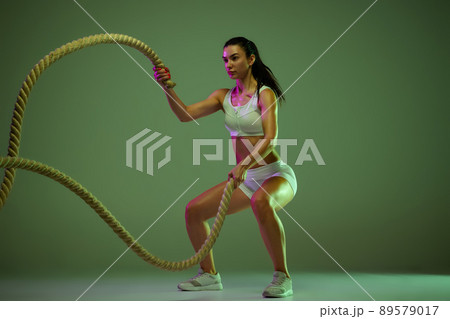 Dynamic portrait of young sportive woman training, doing exercises with rope isolated over green studio background in neon light 89579017