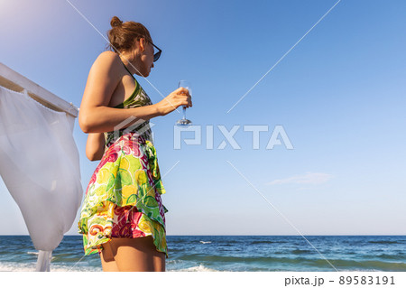 Back view young stylish adult woman holding cold alcohol refresh beverage glass looking at ocean shore at summer seaside vacation. Female person treat relax holiday at exotic resort hotel beach Back view young stylish adult woman holding cold alcohol refresh beverage glass looking at ocean shore at summer seaside vacation. Female person treat relax holiday at exotic resort hotel beach 89583191